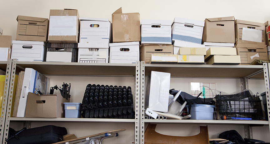 A stack of office supplies piled high in a storage room includes bankers boxes